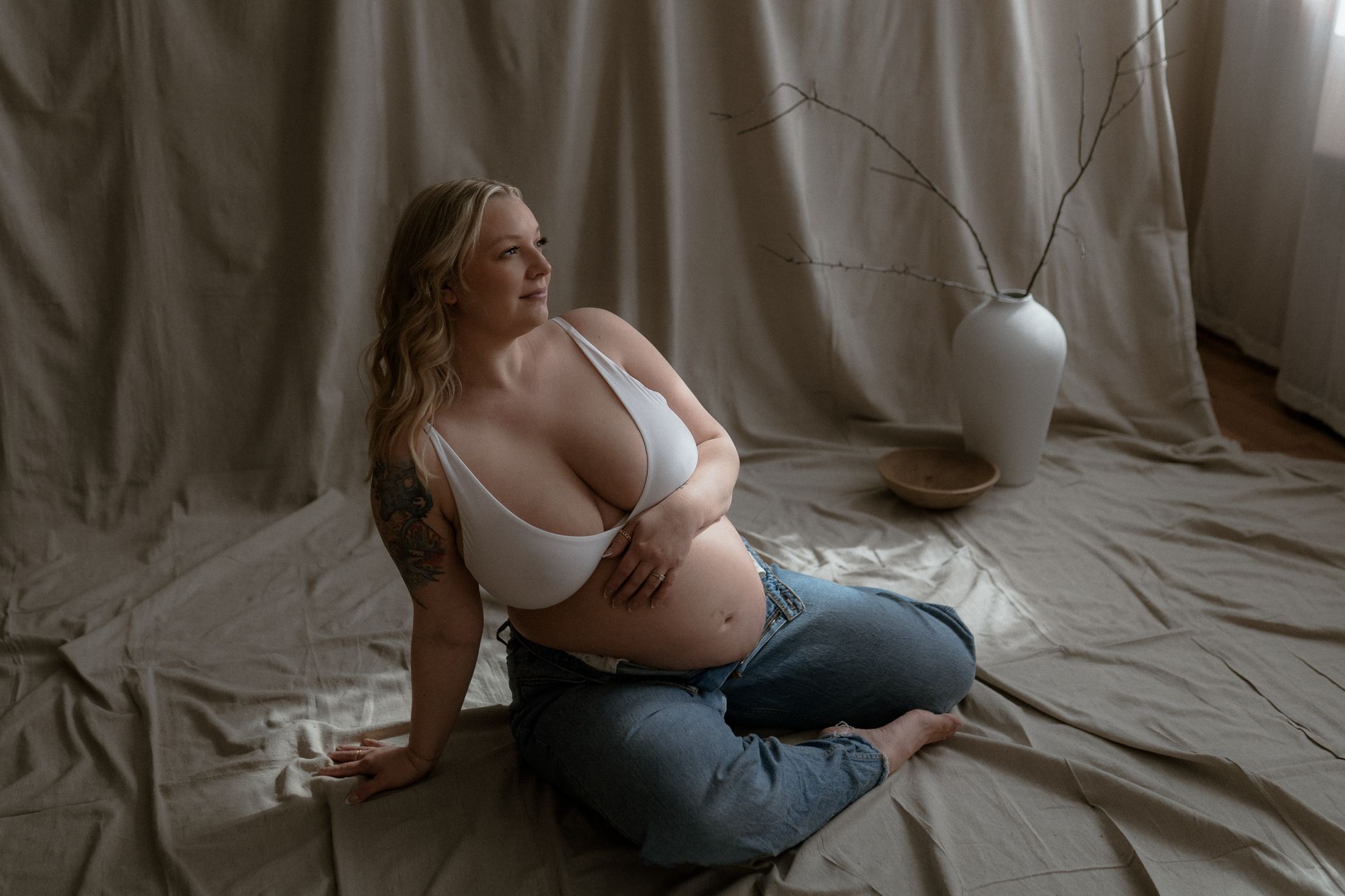 Madison Maternity studio session with a pregnant woman sitting in jeans and a white bra in the sunlight on a draped canvas.
